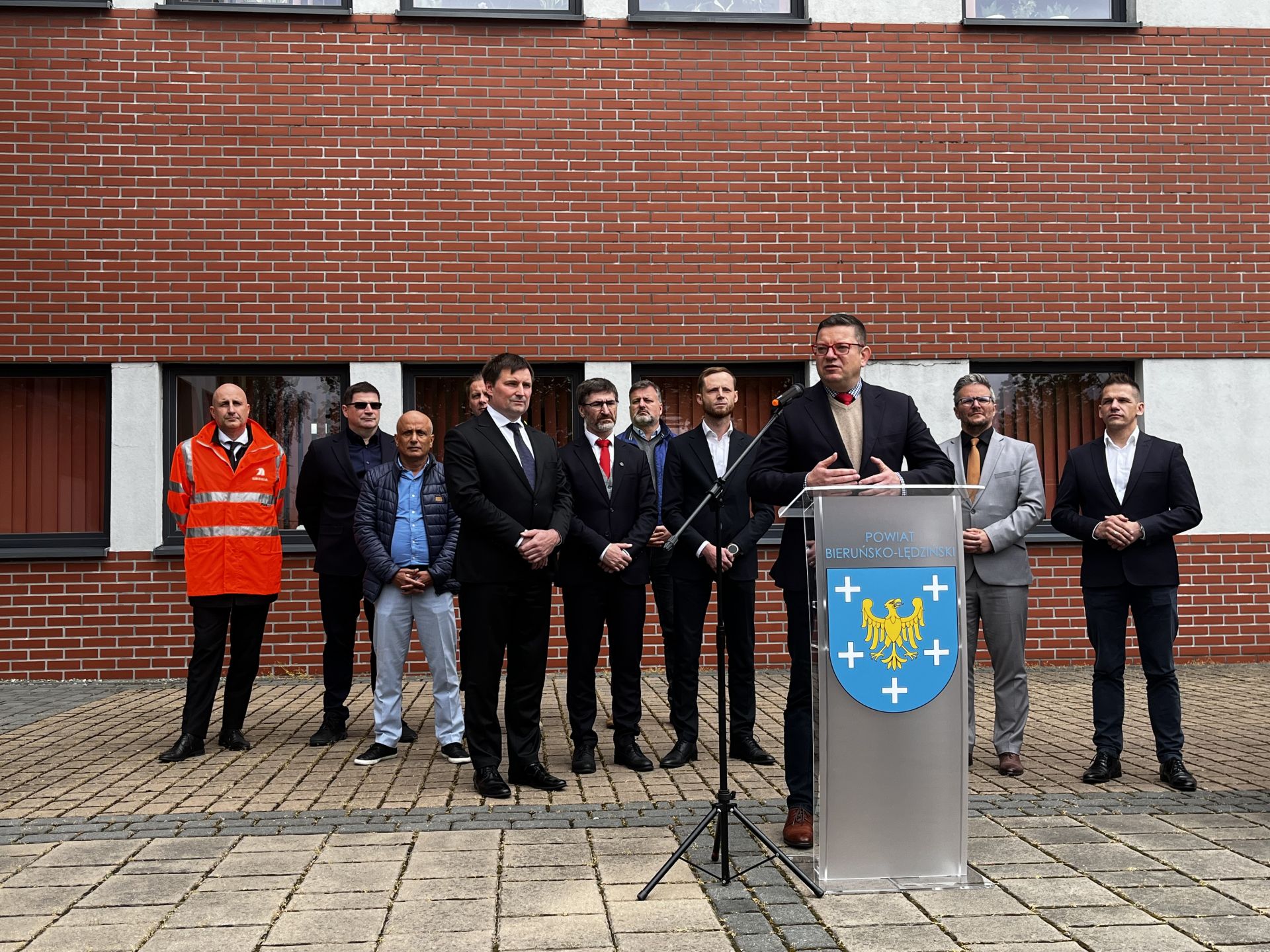 Stojący przed budynkiem z ceglaną elewacją mężczyźni. Jeden z nich przemawia z mówicy z herbem powiatu (na niebieskim tle żółty orzeł wokół niego pięć białych krzyży)